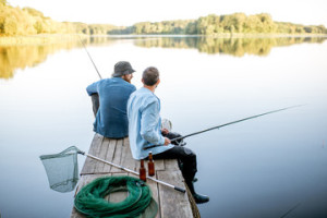 Fototapeta Wędkarstwo Two male friends dressed in blue shirts fishing together with net and rod sitting on the wooden pier during the morning light