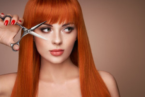 Fototapeta Fryzjerstwo Beautiful young woman with a bright makeup and a smooth long hair holds metal scissors. Model with red hair. Hair salon